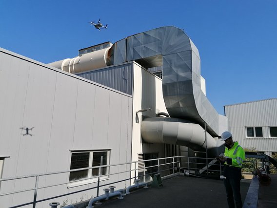 Eine Drohne schwebt über einem modernen Industriegebäude mit auffälliger silberner Architektur und Lüftungsanlagen. Ein Arbeiter in gelber Warnweste steht auf einer Plattform des Gebäudes.