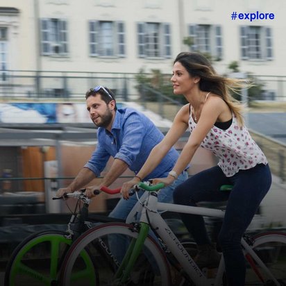 Ein Mann in blauem Hemd und eine Frau in weißem Top mit Blumenmuster fahren gemeinsam Fahrrad entlang einer städtischen Straße. Im Hintergrund ist ein vorbeifahrender Zug und ein klassisches, helles Gebäude mit vielen Fenstern zu sehen.