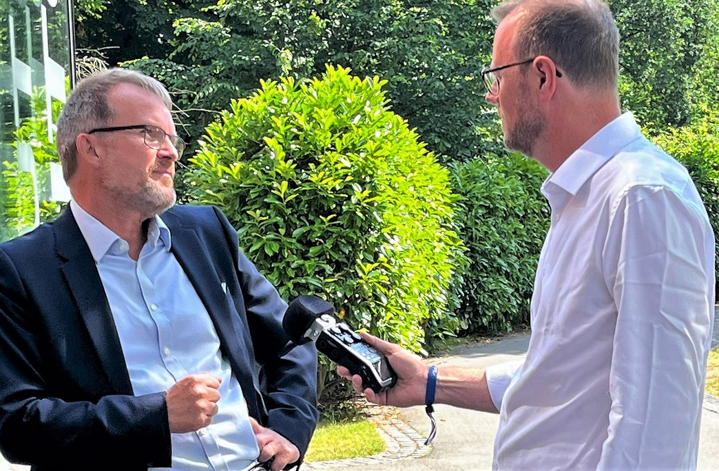 Zwei Männer in Geschäftskleidung im Gespräch im Freien, einer hält ein Audiogerät, vermutlich für ein Interview vor grünem Hintergrund.