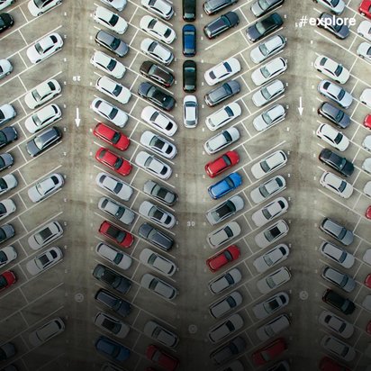 Luftaufnahme eines großen Parkplatzes mit schräg angeordneten Parkbuchten, auf denen überwiegend weiße, schwarze und rote Fahrzeuge in geordneten Reihen parken. Pfeile auf dem Asphalt zeigen die Fahrtrichtung an.