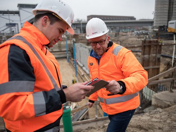 Geologe Matthias und Timo Hackmann in orangefarbenen Sicherheitswesten und Schutzhelmen betrachten gemeinsam Unterlagen auf einer Baustelle.