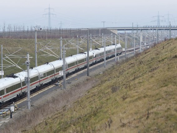  Ein weißer ICE-Hochgeschwindigkeitszug der Deutschen Bahn fährt durch eine ländliche Landschaft mit Oberleitungen und Signalmasten. Im Hintergrund sind Stromleitungen und eine Autobahnbrücke in der flachen, grasbewachsenen Umgebung zu sehen.