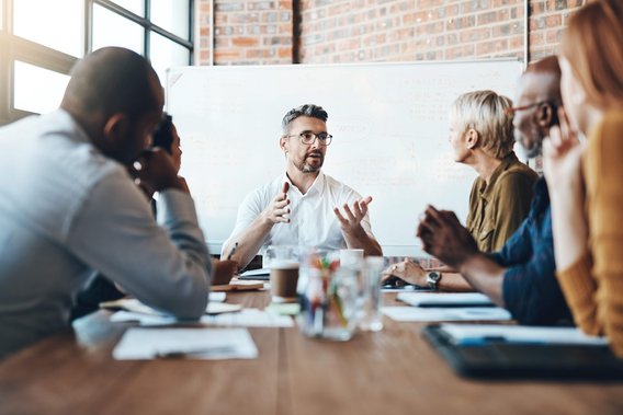 Eine Gruppe von Personen sitzt an einem Konferenztisch und diskutiert vor einem Whiteboard.