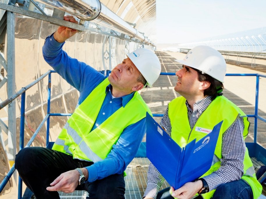 Zwei Männer mit weißen Schutzhelmen und gelben Sicherheitswesten inspizieren Rohrleitungen auf einer erhöhten Plattform. Im Hintergrund sind Teile einer Solaranlage zu erkennen.