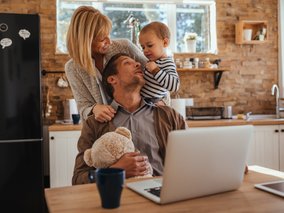 Eine Familie in einer Küche mit Backsteinwand – eine Frau steht hinter einem sitzenden Mann mit Teddybär, während ein Kleinkind mit Streifenshirt seinen Vater berührt.