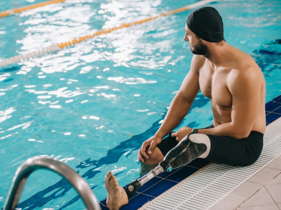 Ein muskulöser Mann mit Badekappe sitzt am Beckenrand eines Schwimmbads und trägt eine Beinprothese.