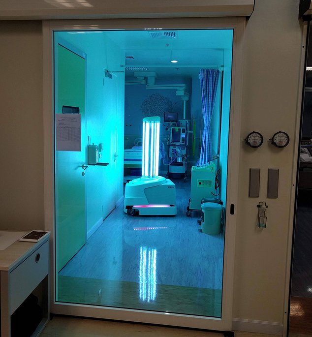 Ein Krankenhauszimmer mit türkisblauer UV-Beleuchtung zur Desinfektion, sichtbar durch eine Glastür mit medizinischen Geräten und einem Patientenbett im Inneren.