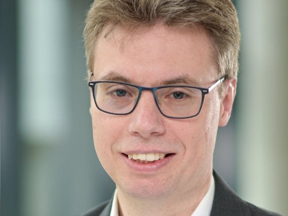 Boris Michael Leidner mit einer blauen Brille lächelt in die Kamera, gekleidet in einem dunklen Sakko vor einem hellen Hintergrund.