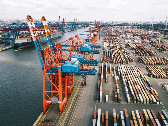 Luftaufnahme eines Containerhafens mit orangefarbenen Kränen, Schiffen und sortierten Containern in Reihen; Logistiksystem mit Windrädern im Hintergrund.