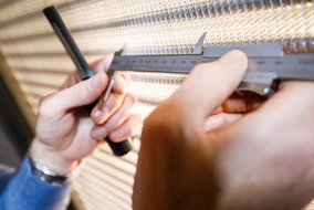 Nahaufnahme von Händen, die mit Messschieber und Stift präzise Messungen an einer strukturierten Metalloberfläche vornehmen.