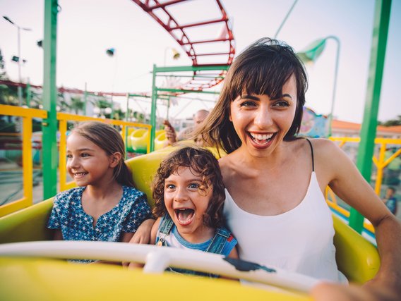 Drei lachende Personen genießen ein gelbes Fahrgeschäft in einem bunten Freizeitpark.