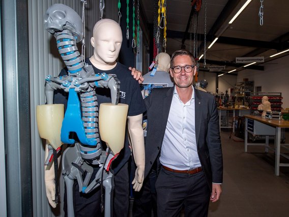 Peter Schimmelpfenning im Anzug steht neben anatomischen Crashtest-Dummies mit technischen Gelenken in einem Testlabor.