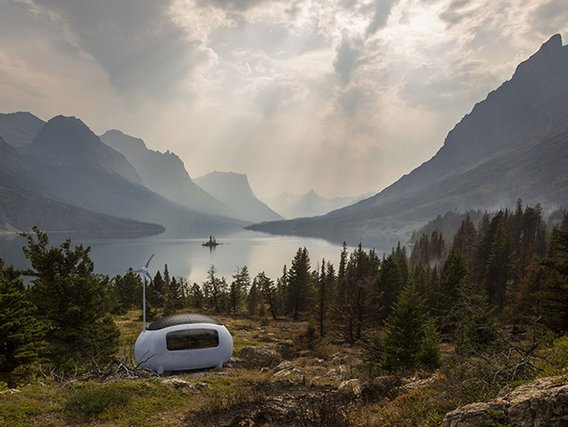 Weißes, futuristisches Mikro-Wohnmodul mit kleinem Windrad steht auf einer Waldlichtung mit Blick auf einen Bergsee in dramatischer Gebirgslandschaft.