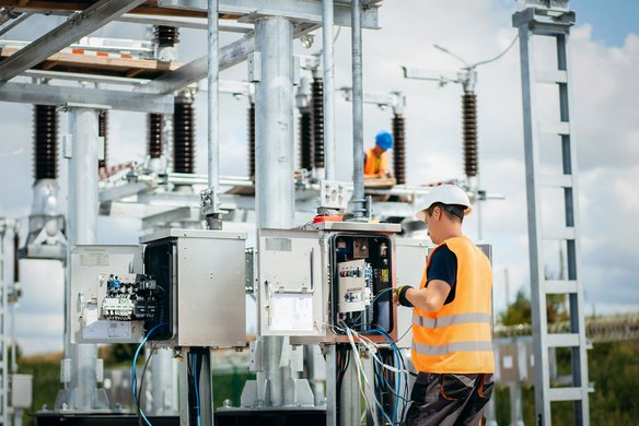 Techniker mit Warnweste und Helm arbeitet an einer offenen Schalttafel in einer Energieanlage im Freien.