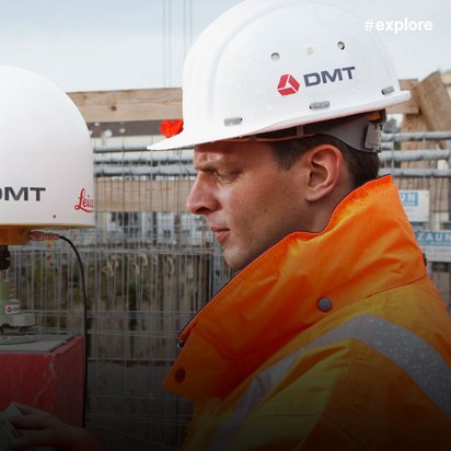 Ein Bauarbeiter in oranger Schutzkleidung und weißem Helm arbeitet mit einem DMT-Vermessungsgerät auf einer aktiven Baustelle mit Kränen im Hintergrund.