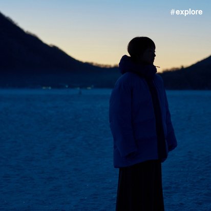 eine Frau steht auf ebenem Boden. Im Hintergrund sind Hügel zu sehen, und der Himmel ist dunkel. Hinter den Hügeln geht die Sonne unter.