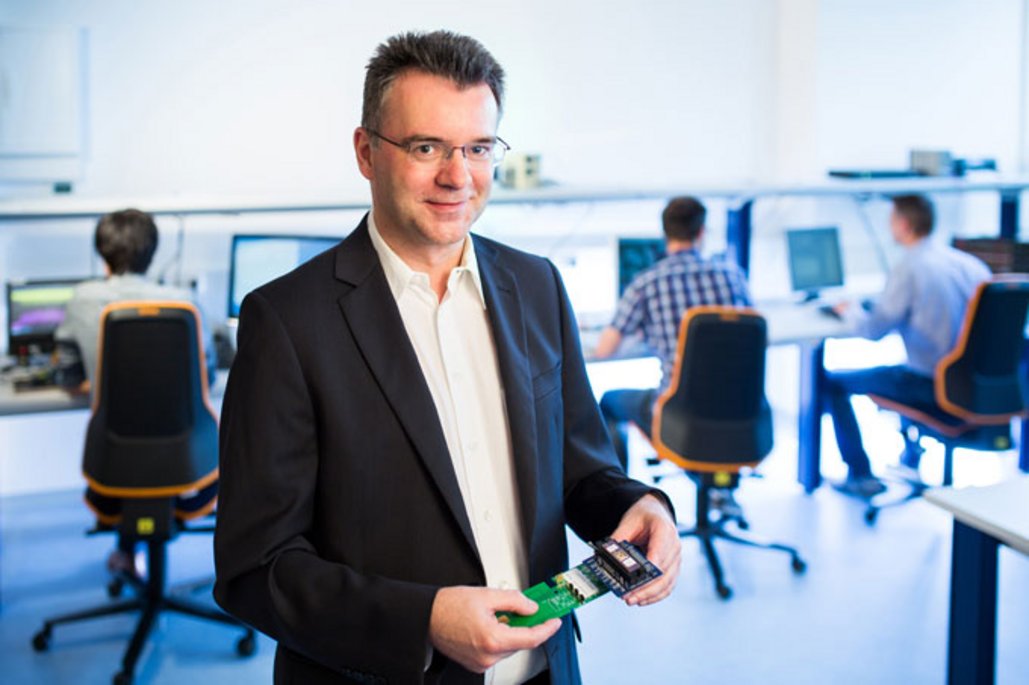 Patrick Bödeker in dunklem Anzug und Brille hält eine grüne Platine und lächelt in einem modernen Büro mit Arbeitsplätzen im Hintergrund.