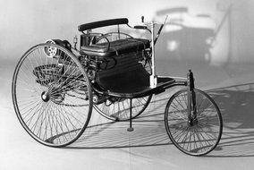 Schwarz-weiß Foto des ersten Automobils von Carl Benz mit drei Speichenrädern und dampfbetriebener Technik.