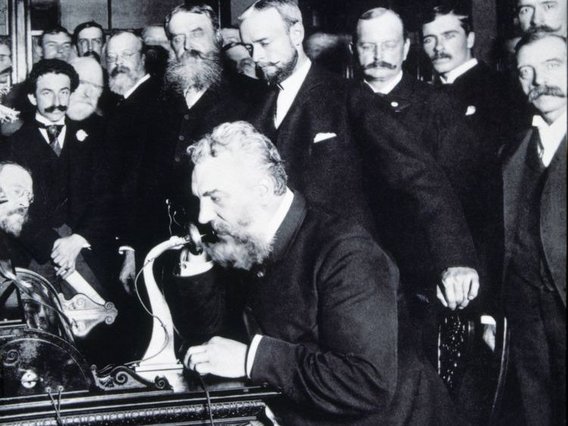 Eine historische Schwarzweißfotografie zeigt Alexander Graham Bell beim ersten Ferntelefongespräch 1892. Er sitzt an einem Schreibtisch mit einem frühen Telefongerät, umgeben von mehreren Männern in formeller viktorianischer Kleidung mit charakteristischen Bärten und Schnurrbärten.