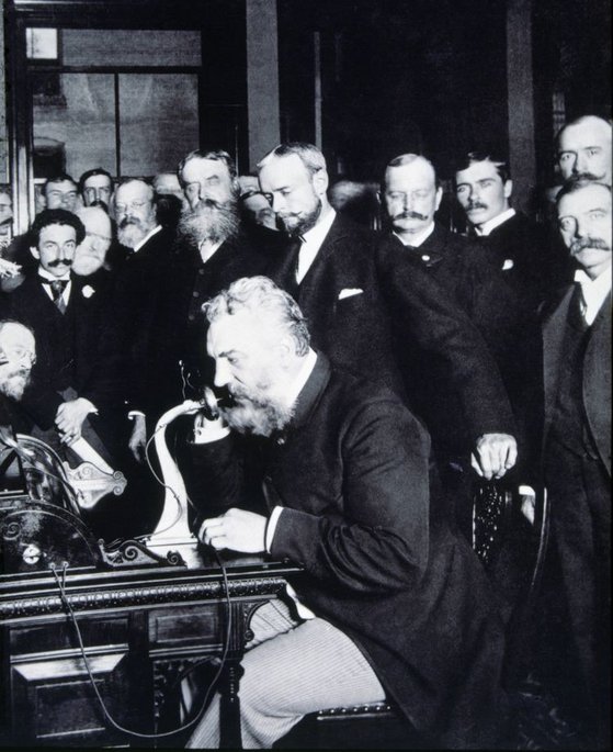 Eine historische Schwarzweißfotografie zeigt Alexander Graham Bell beim ersten Ferntelefongespräch 1892. Er sitzt an einem Schreibtisch mit einem frühen Telefongerät, umgeben von mehreren Männern in formeller viktorianischer Kleidung mit charakteristischen Bärten und Schnurrbärten.