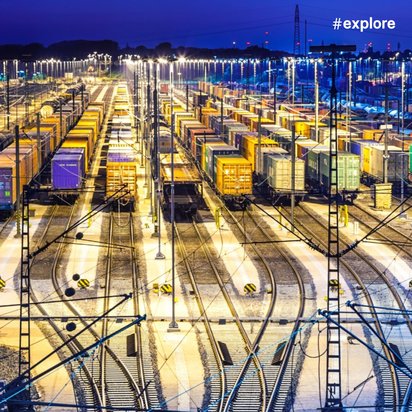 Ein beleuchteter Güterbahnhof bei Nacht mit zahlreichen farbigen Containern auf parallelen Gleisen. Die Szene ist in blauem und gelbem Licht getaucht.