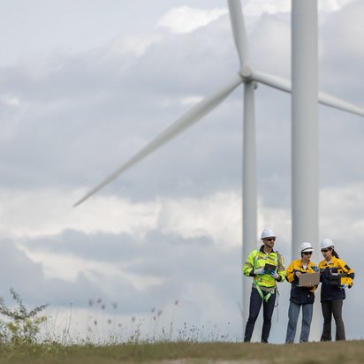 Drei Fachkräfte mit Schutzkleidung stehen vor einem großen Windrad und prüfen etwas auf einem Laptop.