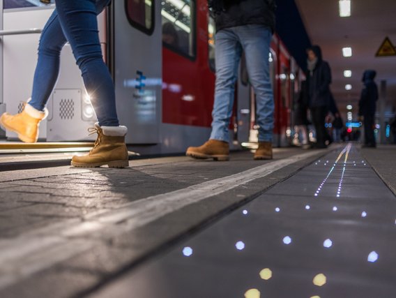 Fahrgäste steigen aus einem rot-weißen Zug an einem nächtlichen Bahnsteig aus, wobei LED-Lichtstreifen im Boden zur Navigation leuchten. Die Szene zeigt moderne Bahnhofsbeleuchtung und Sicherheitstechnik für bessere Orientierung bei Dunkelheit.