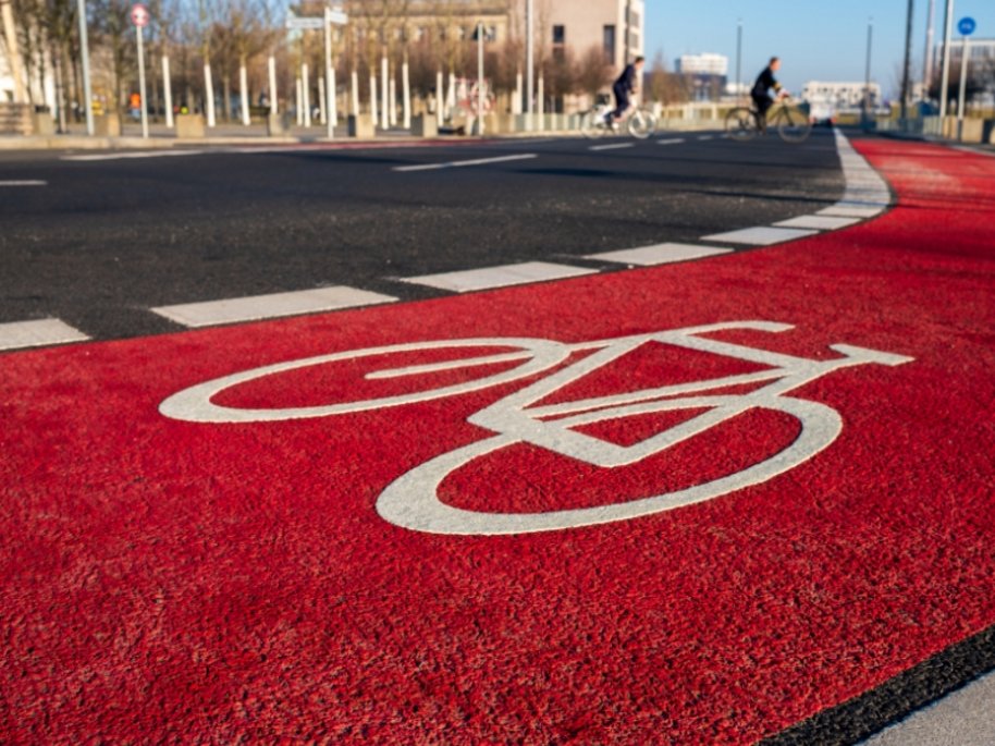 Roter Radweg mit weißem Fahrrad-Symbol und Markierungsstreifen, Radfahrer im unscharfen Hintergrund auf Stadtstraße.