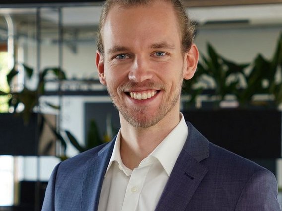 A professional portrait of Patrick Krieger with light brown hair tied back. He is wearing a navy blue suit and a white shirt. The background is a modern office with plants and glass partitions, creating a professional yet welcoming atmosphere.