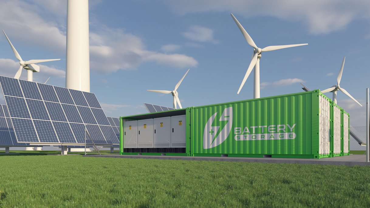A green battery storage system stands on a meadow, surrounded by solar panels and wind turbines. The solar panels are arranged in rows and the wind turbines rotate in the background under a blue sky with a few clouds.