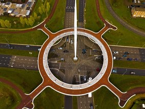Luftaufnahme eines großen Kreisverkehrs mit weißem Mast in der Mitte, umgeben von braunen Radwegen und grünen Flächen.