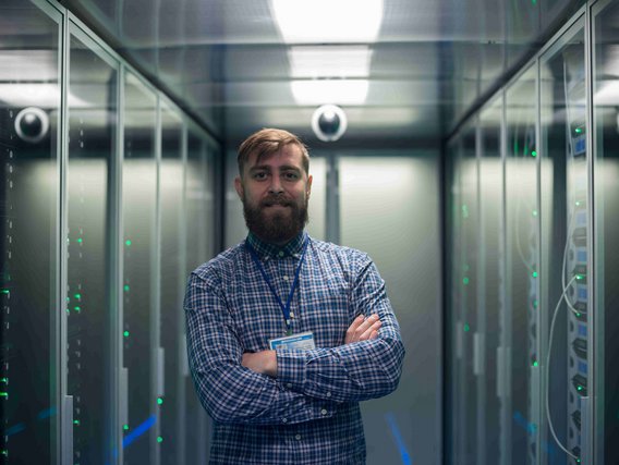 Person steht mit verschränkten Armen in einem Serverraum zwischen zwei Reihen von Server-Racks.