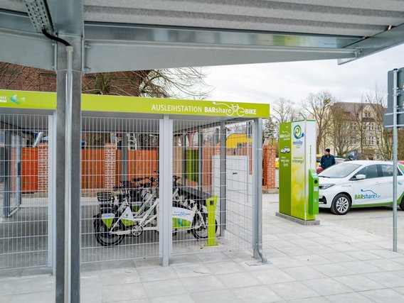 Eine überdachte Fahrradverleihstation mit grünen "BARshare BIKE" Schildern, in der mehrere Leihfahrräder in einem vergitterten Bereich stehen. Im Hintergrund sind Elektroautos und Gebäude zu sehen.