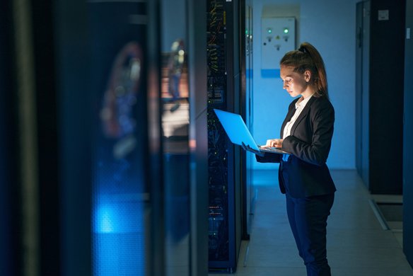 Ein Frau mit einem Laptop in den Händen steht in einem Rechenzentrum. Sie schaut auf den Bildschirm des Laptops.