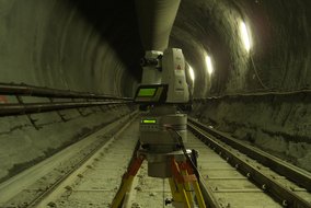 Ein weißes Leica-Vermessungsgerät steht autonom auf gelbem Stativ in einem Tunnel. Das Gerät mit grünem Display führt automatische Messungen zwischen den Bahngleisen durch.