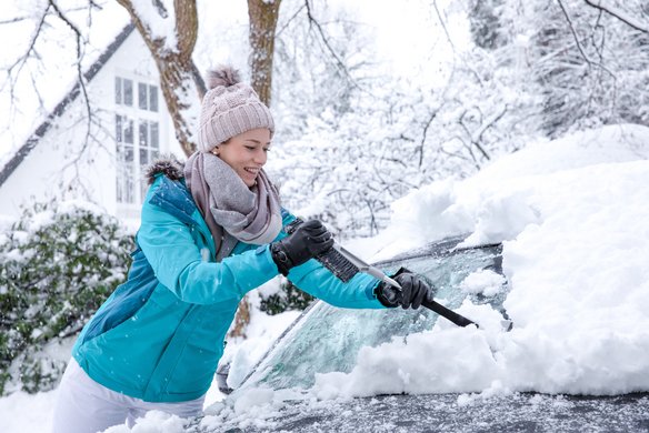 Person in Winterkleidung räumt Schnee von einem Auto in einer verschneiten Umgebung.