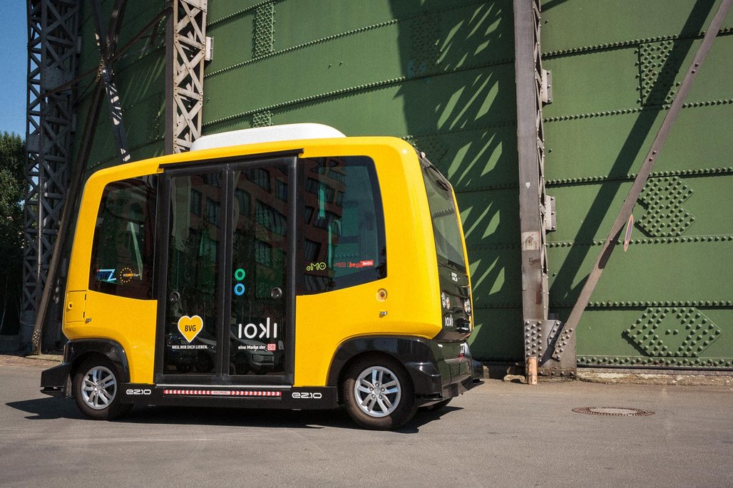  Ein gelber autonomer Kleinbus der BVG steht vor einer grünen Stahlbrücke auf einer Straße.
