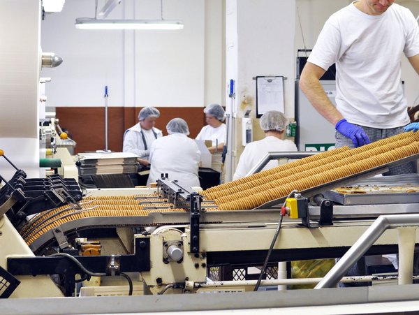 Mitarbeitende an einem Laufband der Keksherstellung in weißen Shirts und mit Hauben