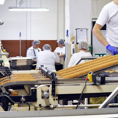 Mitarbeitende an einem Laufband der Keksherstellung in weißen Shirts und mit Hauben