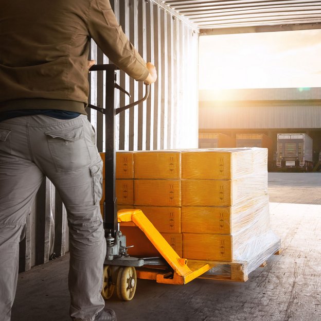 Arbeiter bewegt mit Hubwagen eine Palette mit Kartons aus einem Container in Richtung Laderampe bei Sonnenlicht.