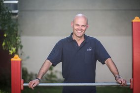 Ein lächelnder Mann in dunklem Poloshirt steht zwischen orangefarbenen und roten Spielgerätelementen im Freien. Er wirkt zufrieden und entspannt.