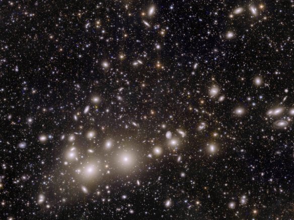 Eine astronomische Aufnahme eines Galaxienhaufens gegen den schwarzen Weltraum. Zahlreiche elliptische und runde Galaxien erscheinen als weiße und gelbliche Lichtflecken unterschiedlicher Größe.