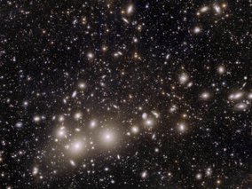 Eine astronomische Aufnahme eines Galaxienhaufens gegen den schwarzen Weltraum. Zahlreiche elliptische und runde Galaxien erscheinen als weiße und gelbliche Lichtflecken unterschiedlicher Größe.