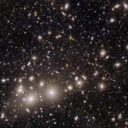 Eine astronomische Aufnahme eines Galaxienhaufens gegen den schwarzen Weltraum. Zahlreiche elliptische und runde Galaxien erscheinen als weiße und gelbliche Lichtflecken unterschiedlicher Größe.