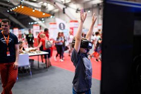 Ein Kind mit VR-Brille steht mit erhobenen Armen in einem Erlebnisbereich. Im Hintergrund ist ein Messehallenboden mit rotem Teppich sowie andere Besucher zu sehen.