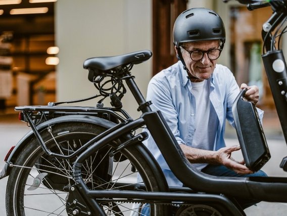 Ein älterer Mann mit Brille und Helm untersucht ein modernes E-Bike in einem Innenraum, wobei er die Batterie herausnimmt.