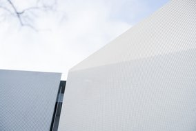 Nahaufnahme einer strukturierten weißen Fassade eines modernen Gebäudes bei Tageslicht. Im Hintergrund ist der Himmel und ein Teil einer weiteren Gebäudefassade sichtbar.