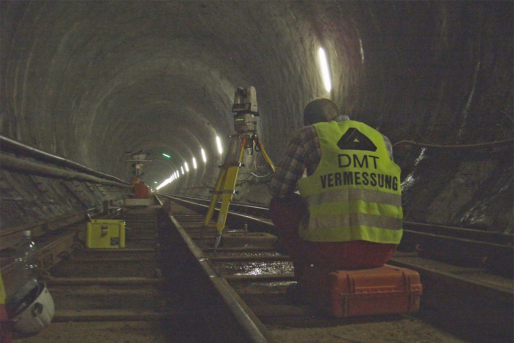 Ein Vermessungstechniker in gelber Warnweste mit "DMT VERMESSUNG" Aufdruck arbeitet in einem beleuchteten Tunnel. Er positioniert ein Vermessungsgerät auf gelbem Stativ zwischen den Gleisen.