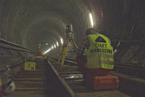 Ein Vermessungstechniker in gelber Warnweste mit "DMT VERMESSUNG" Aufdruck arbeitet in einem beleuchteten Tunnel. Er positioniert ein Vermessungsgerät auf gelbem Stativ zwischen den Gleisen.