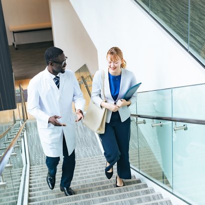 Qualitätsmanagementbeauftragter– Gesundheitswesen Eine Person in einem Kittel und eine Person mit einem Tablet gehen eine Treppe hoch und unterhalten sich.